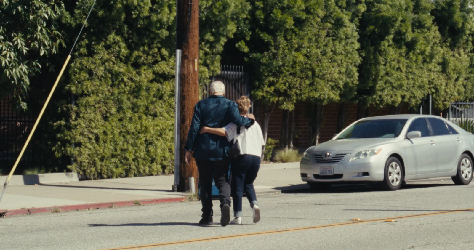 Two people walking arm in arm down a sunlit street past a silver car and a tall hedge, with M3-Film at its default settings.